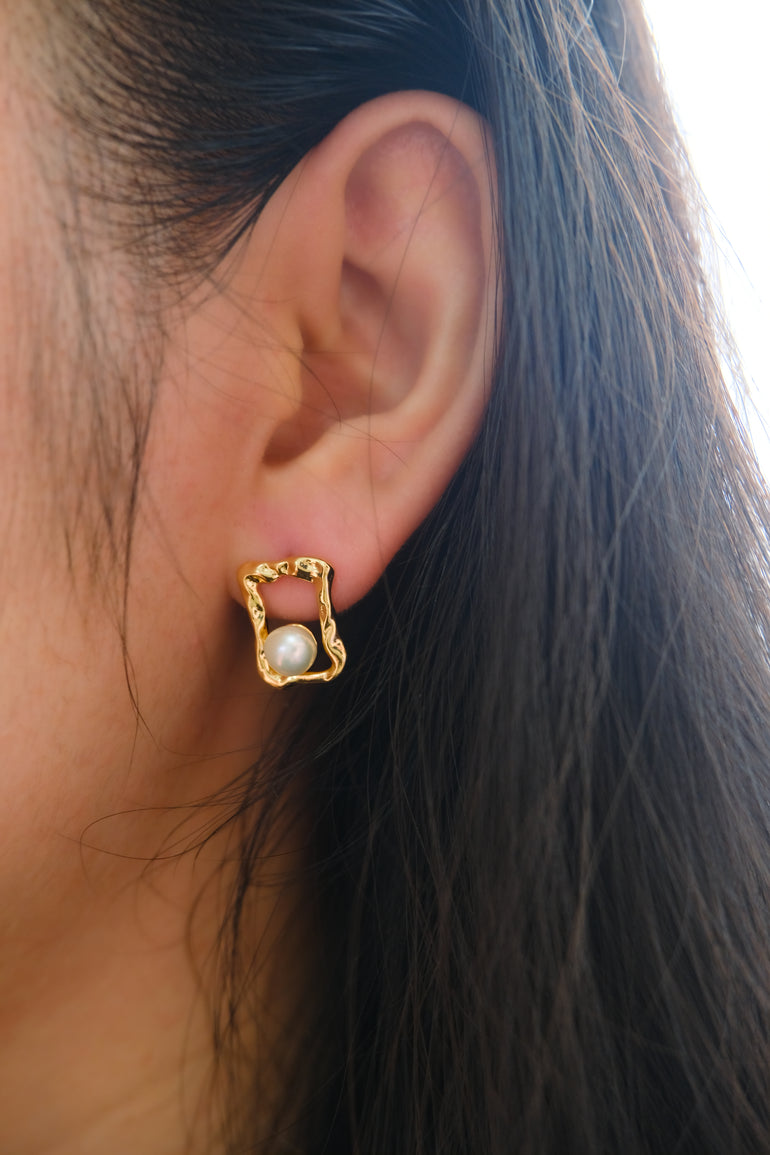 A gold quadrate stud earring with a freshwater pearl on a person's ear.