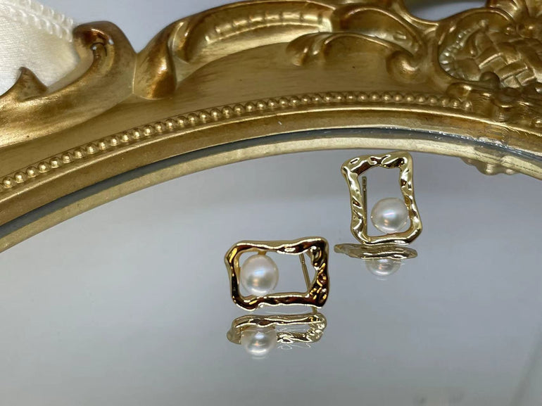 Gold quadrate stud earrings with a freshwater pearl, displayed on a mirrored surface.