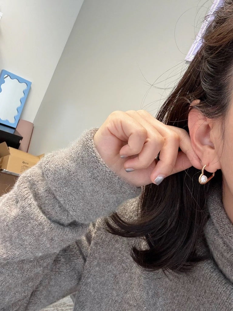 A person wearing a gray sweater holds a heart-shaped earring with a freshwater pearl.