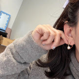 A person wearing a gray sweater holds a heart-shaped earring with a freshwater pearl.