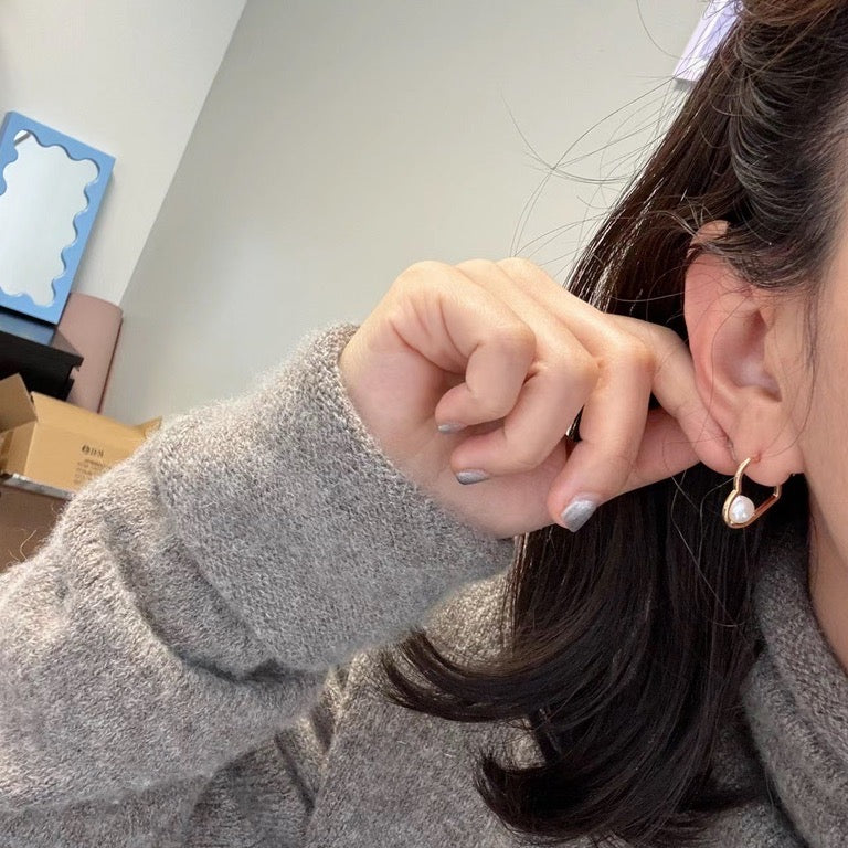 A person wearing a gray sweater holds a heart-shaped earring with a freshwater pearl.