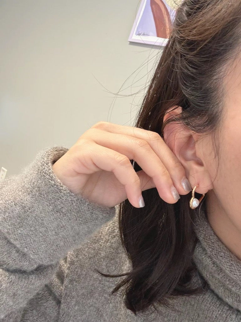 A woman wears a gray sweater and a gold heart earring with a freshwater pearl.