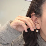 A woman wears a gray sweater and a gold heart earring with a freshwater pearl.
