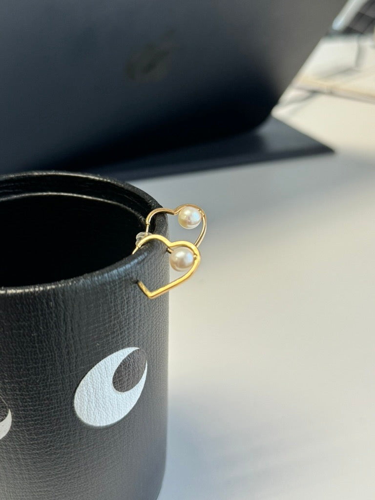 Gold heart-shaped earrings with freshwater pearls resting on a black leather cup.