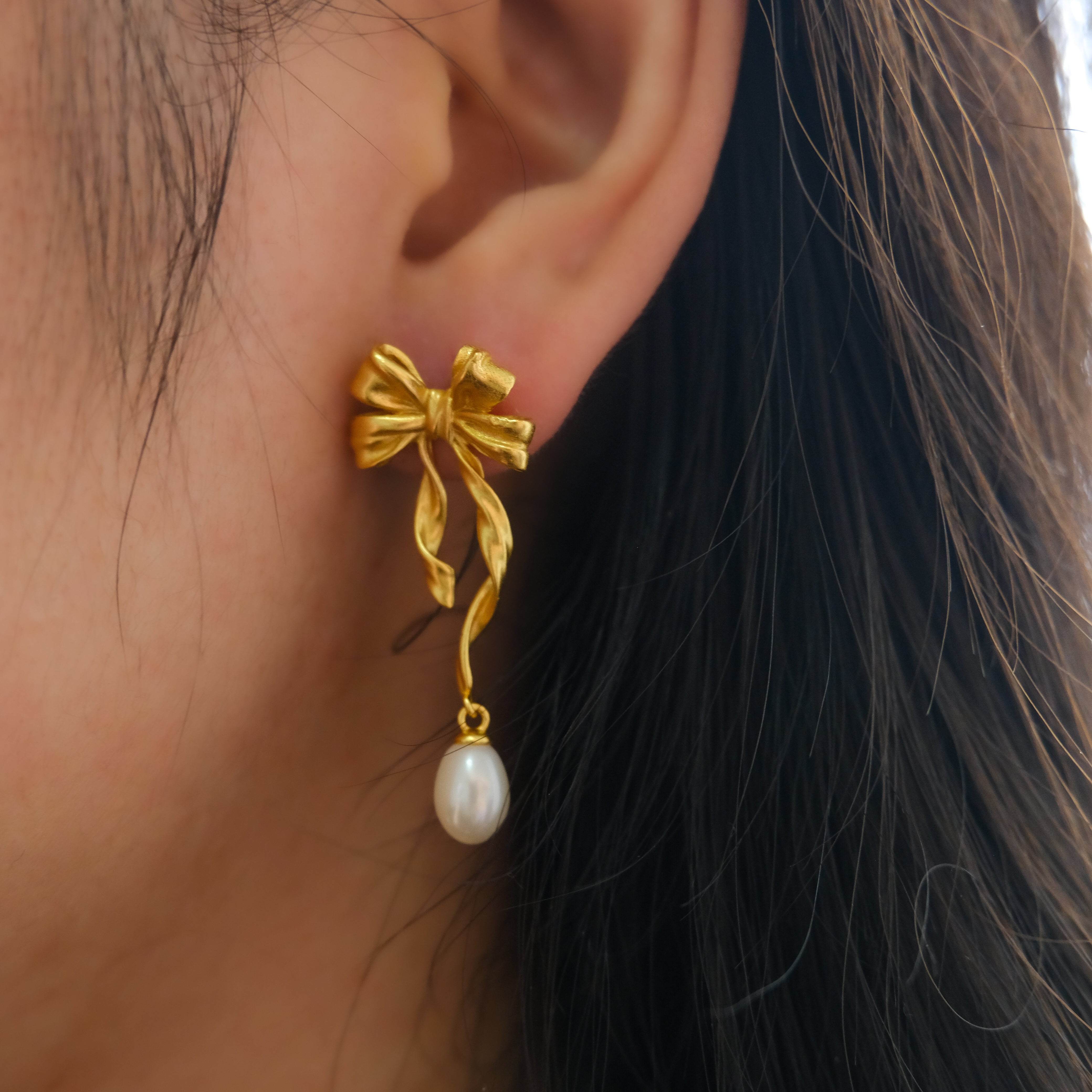 Gold bow earrings with a dangling freshwater pearl on a woman's ear.