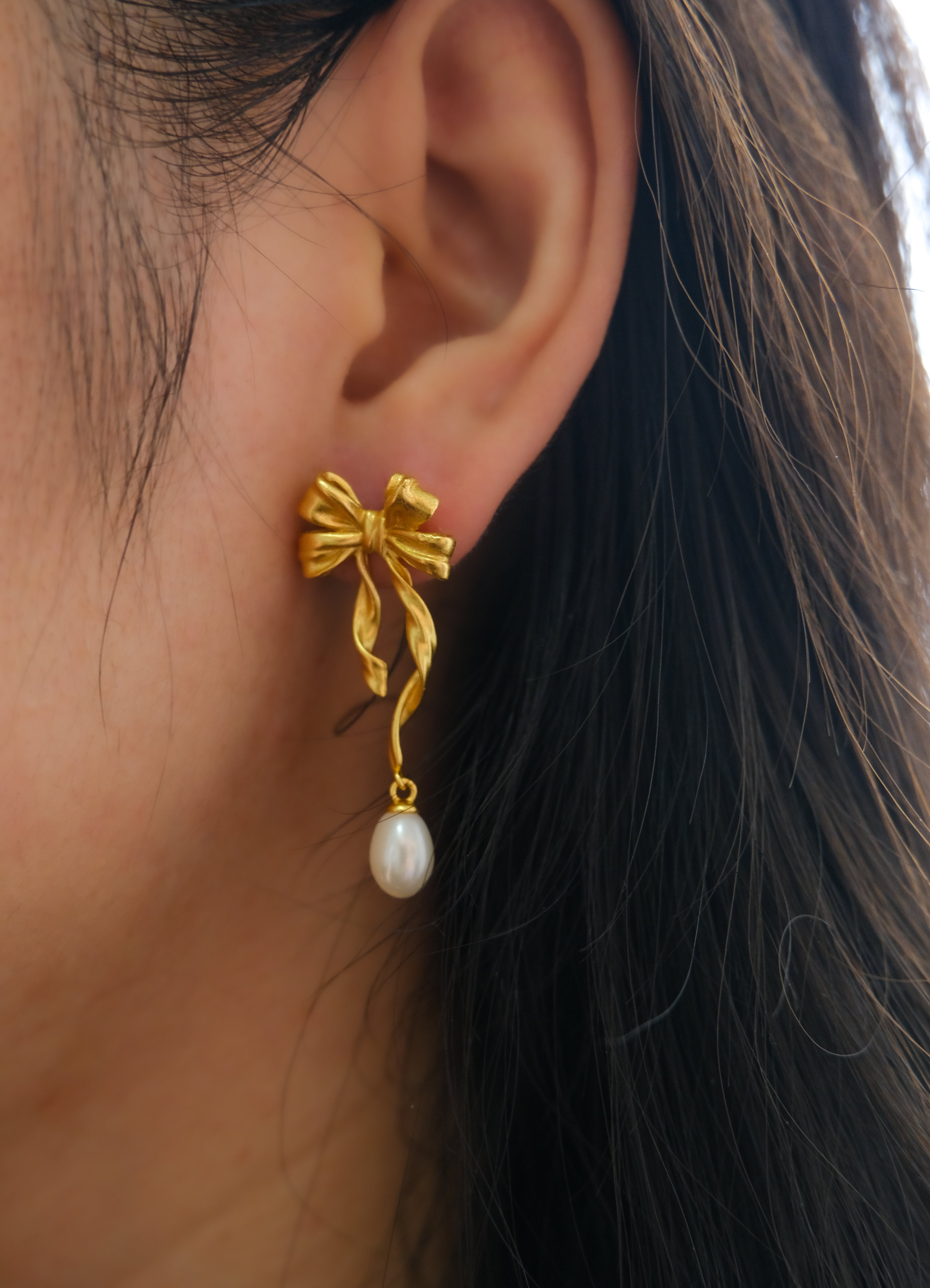 Gold bow earrings with a dangling freshwater pearl on a woman's ear.
