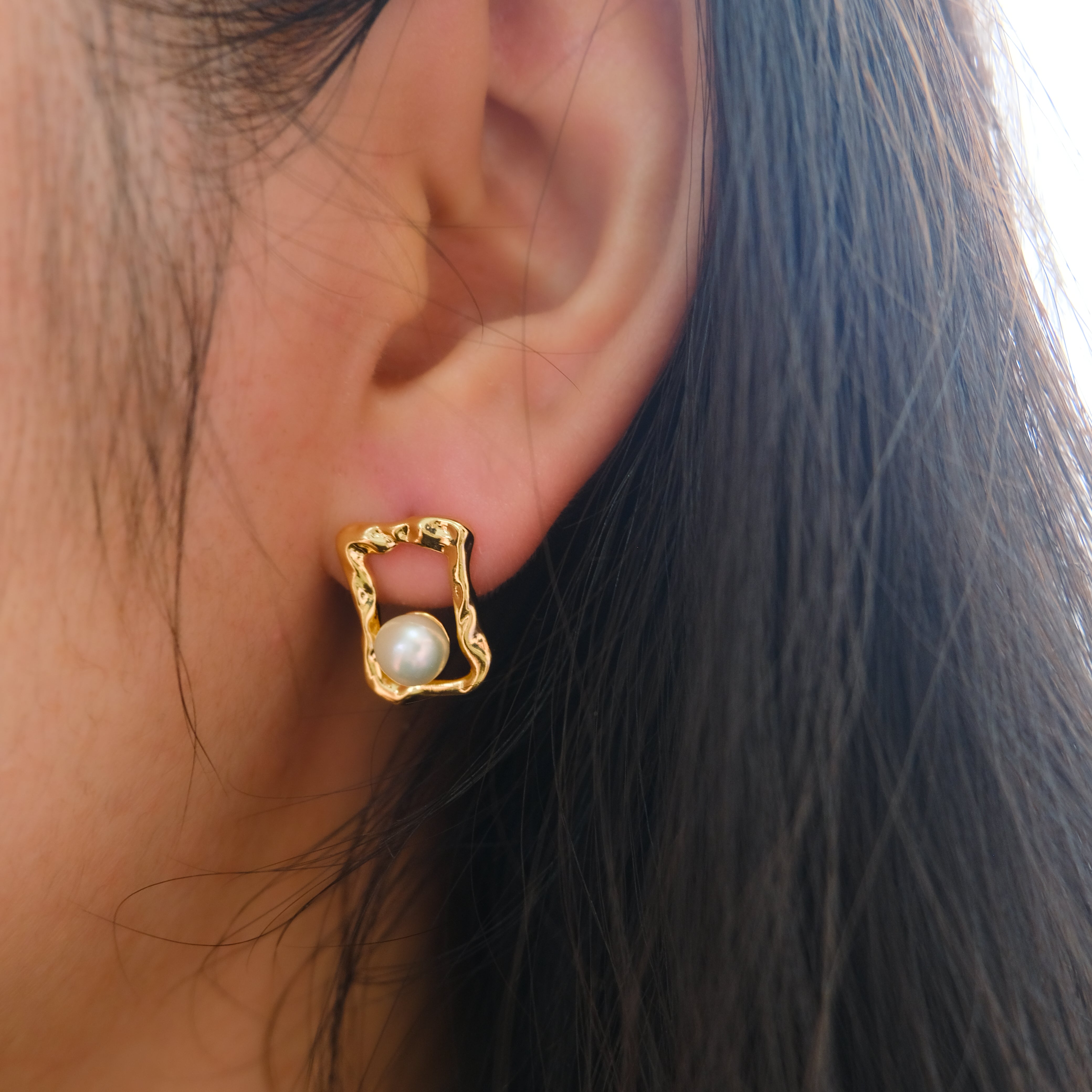 A gold quadrate stud earring with a freshwater pearl on a person's ear.
