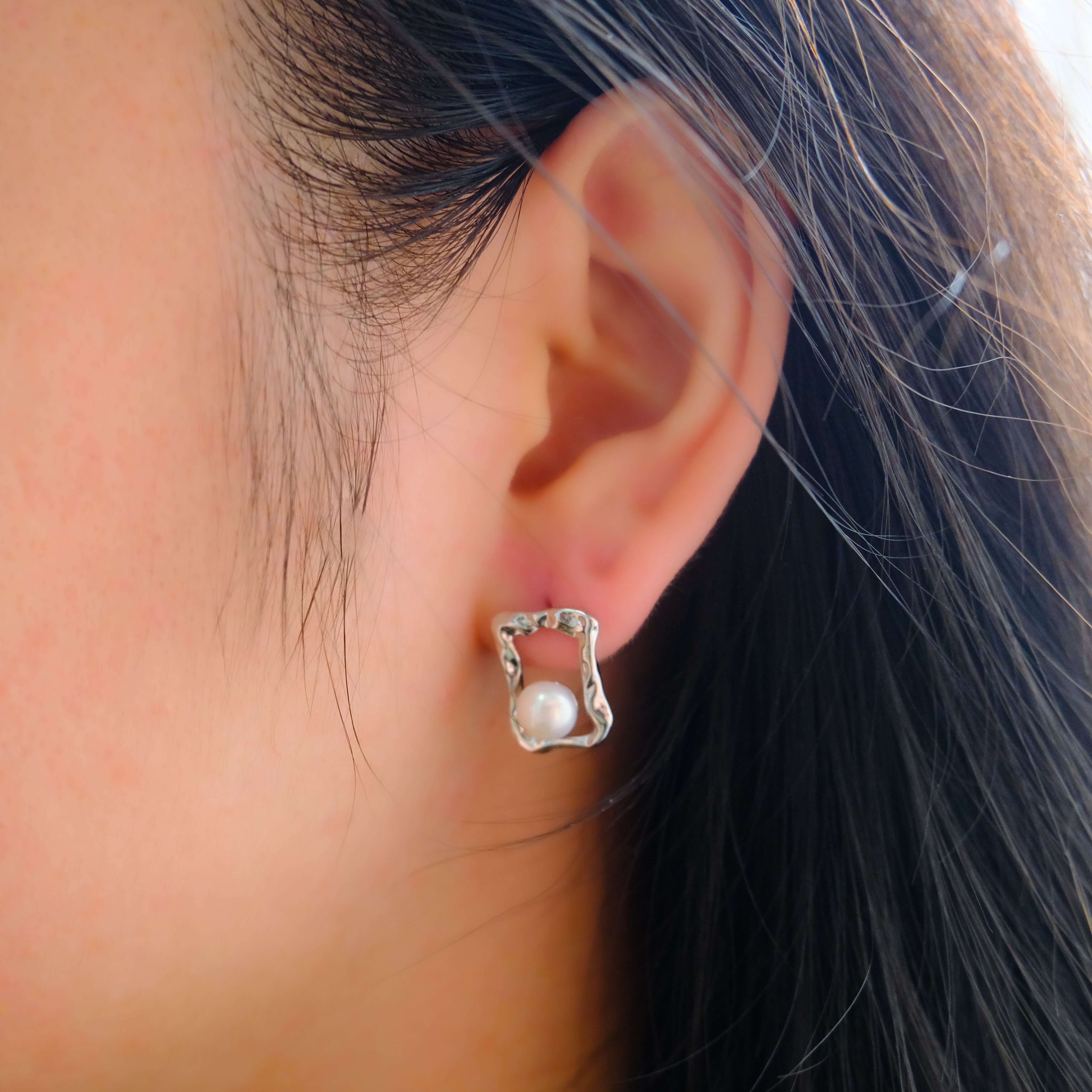 A silver quadrate stud earring with a freshwater pearl on a person's ear.
