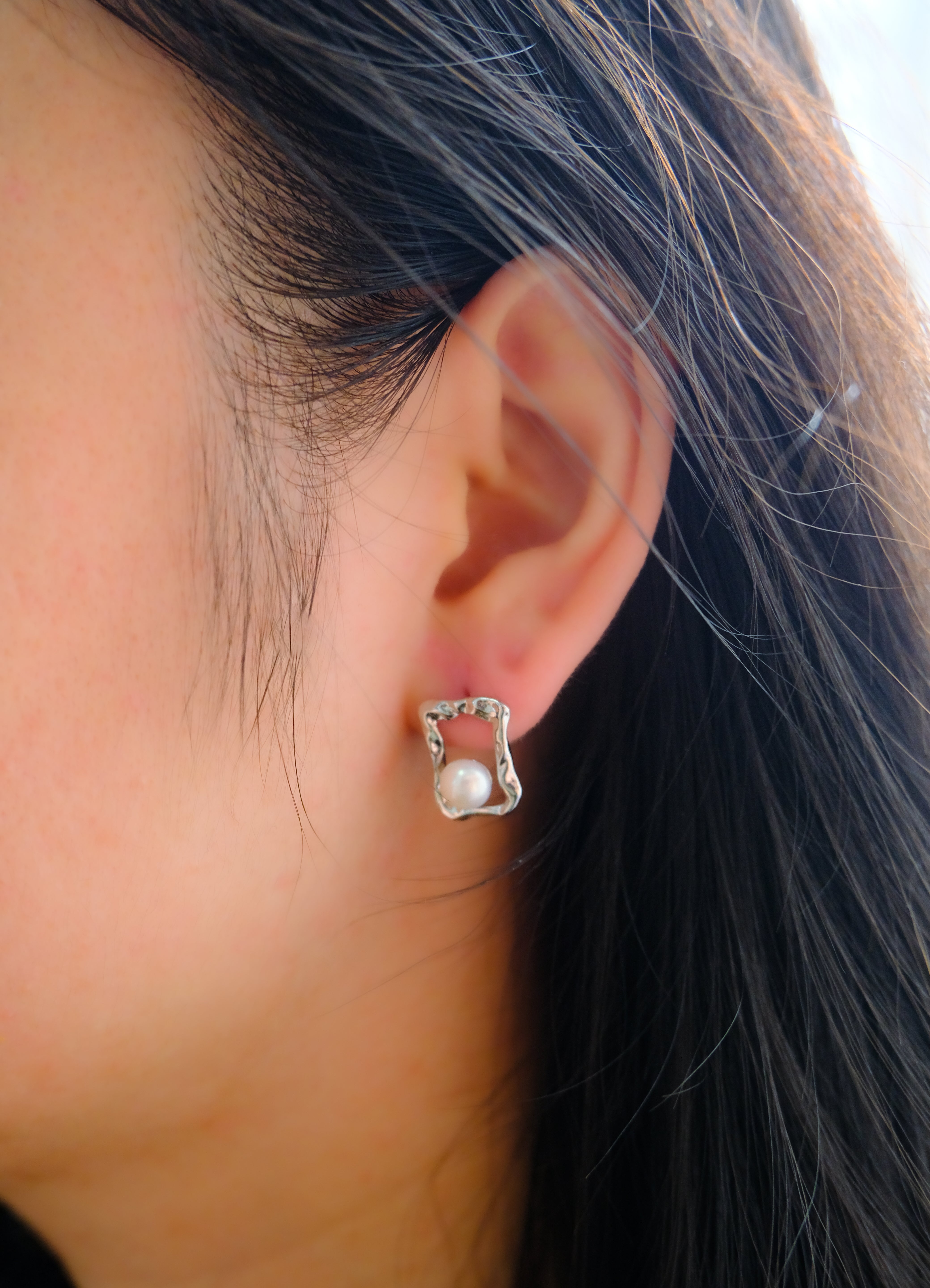 A silver quadrate stud earring with a freshwater pearl on a person's ear.