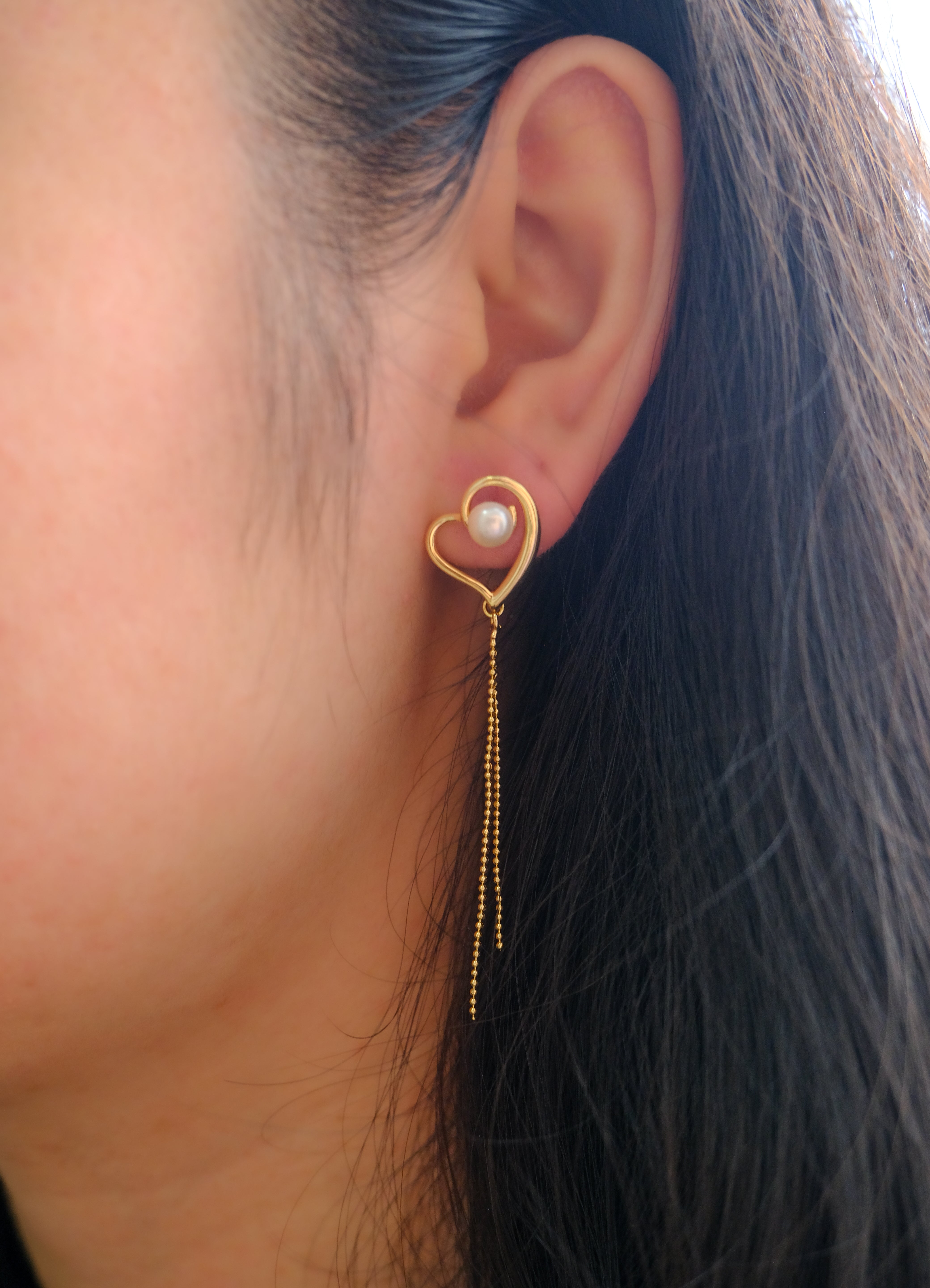 A gold heart-shaped earring with a pearl and dangling chains on a woman's ear.