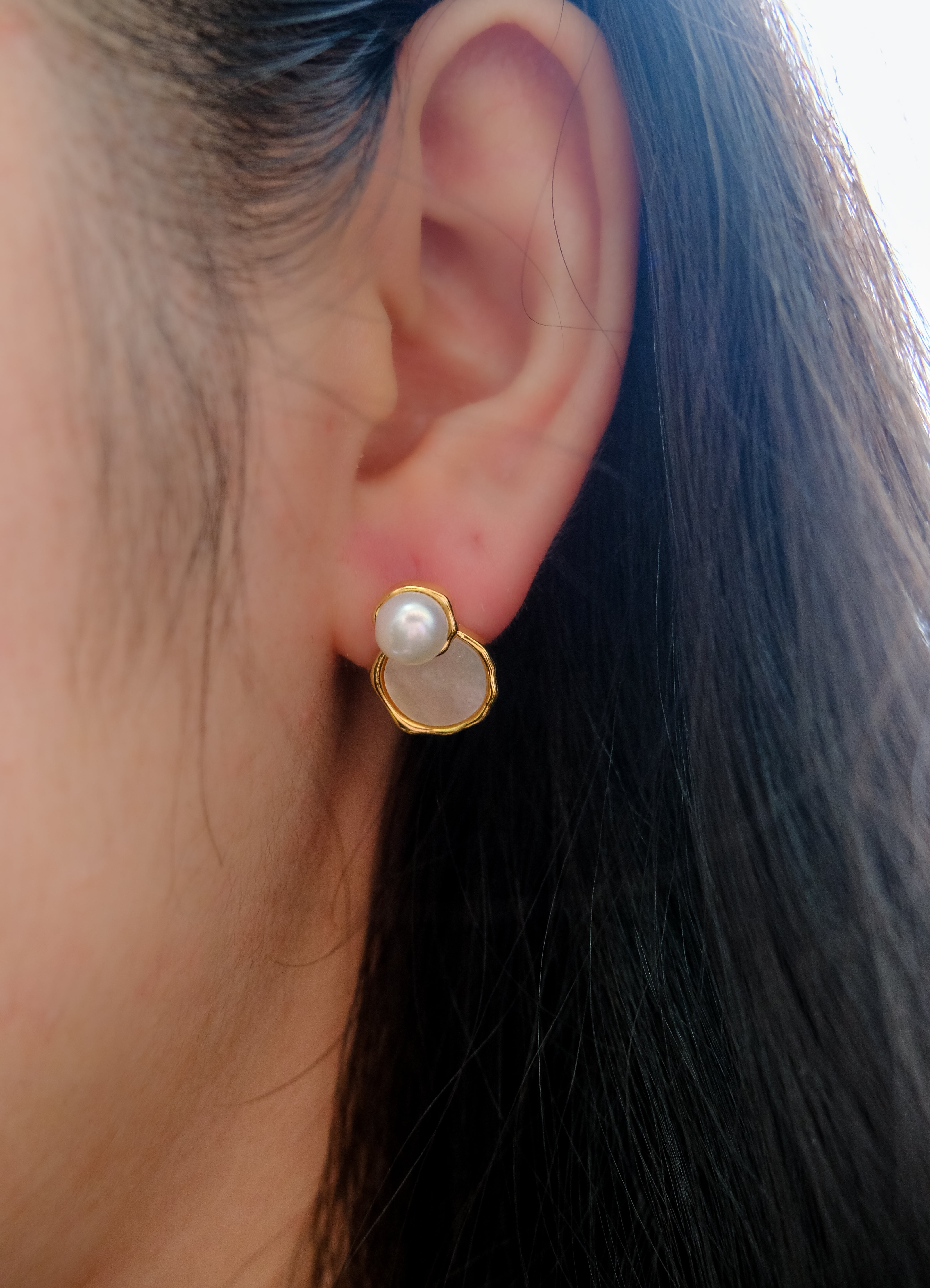 A close-up of a woman's ear wearing a gold earring with a pearl and a white stone.