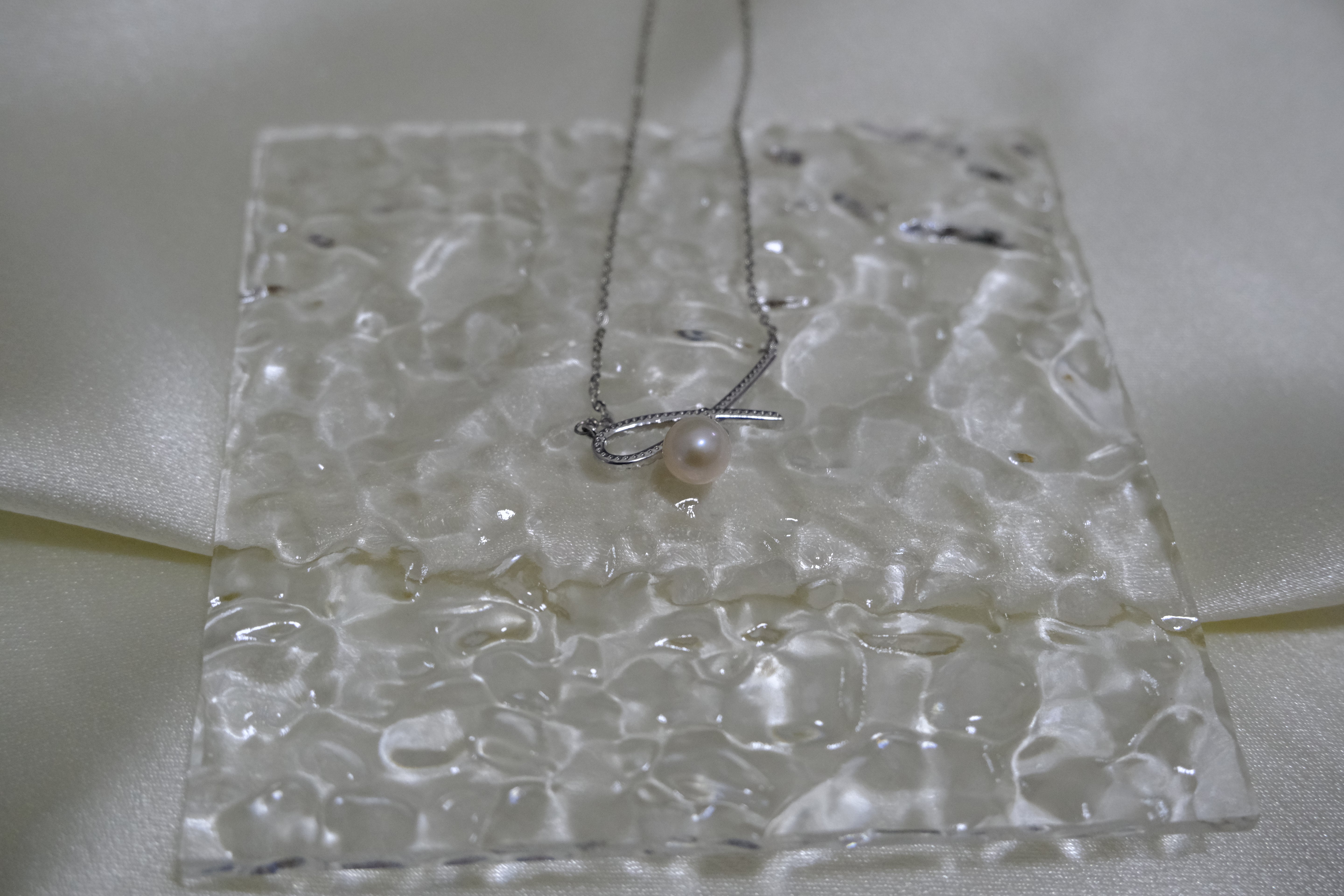 A silver bow pendant necklace with a single freshwater pearl on a textured white surface.