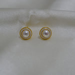 Round freshwater pearl earrings with gold-plated circular frames on a white fabric.