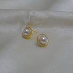 Two round freshwater pearl earrings with gold-plated frames rest on a white silk fabric.