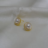 Two round freshwater pearl earrings with gold-plated frames rest on a white silk fabric.
