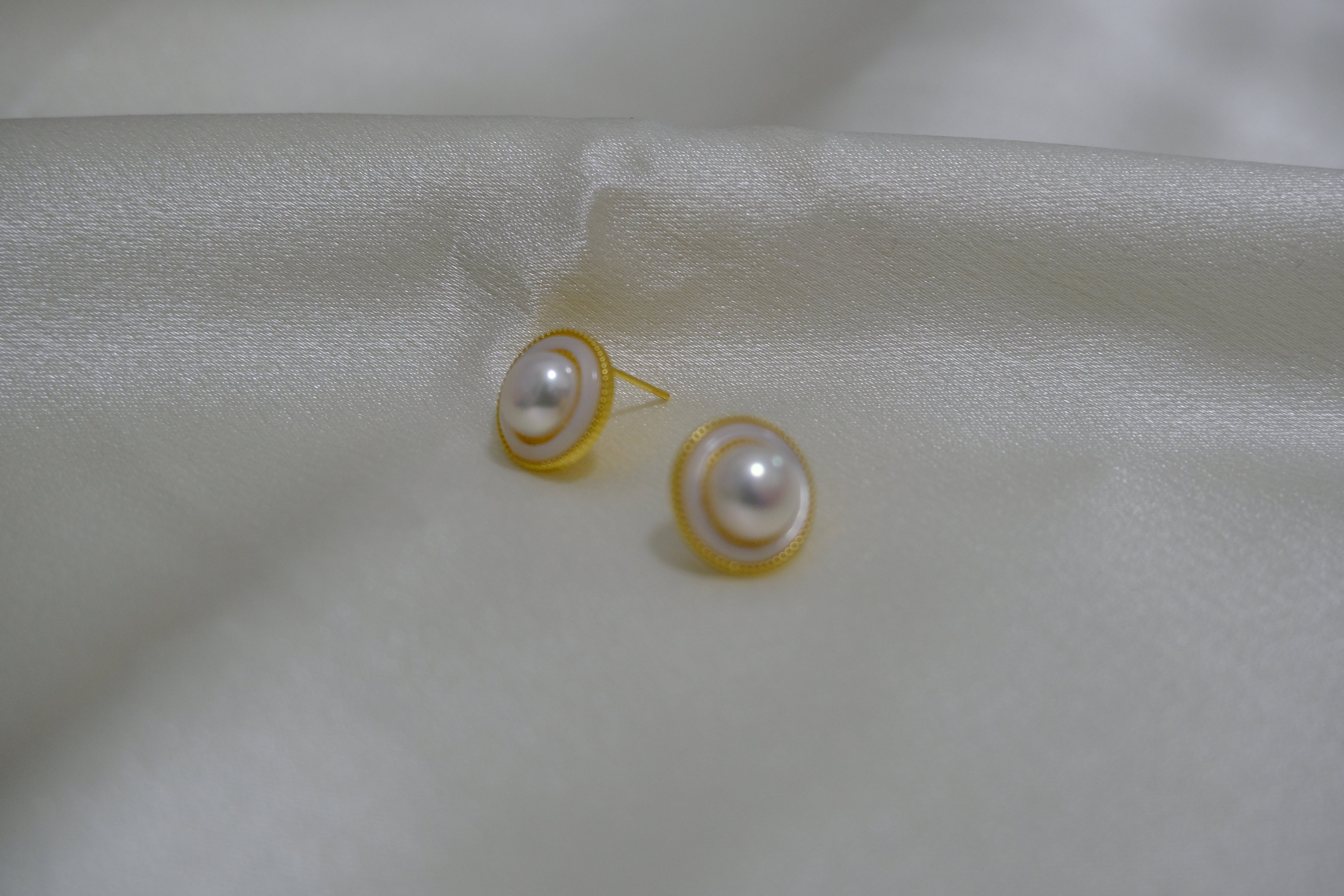 Two round freshwater pearl earrings with gold-plated frames rest on a white silk fabric.
