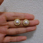 Two round freshwater pearl earrings with gold-plated frames are held in a hand.