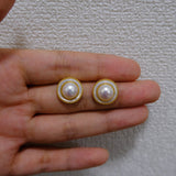 Two round freshwater pearl earrings with gold-plated frames are held in a hand.
