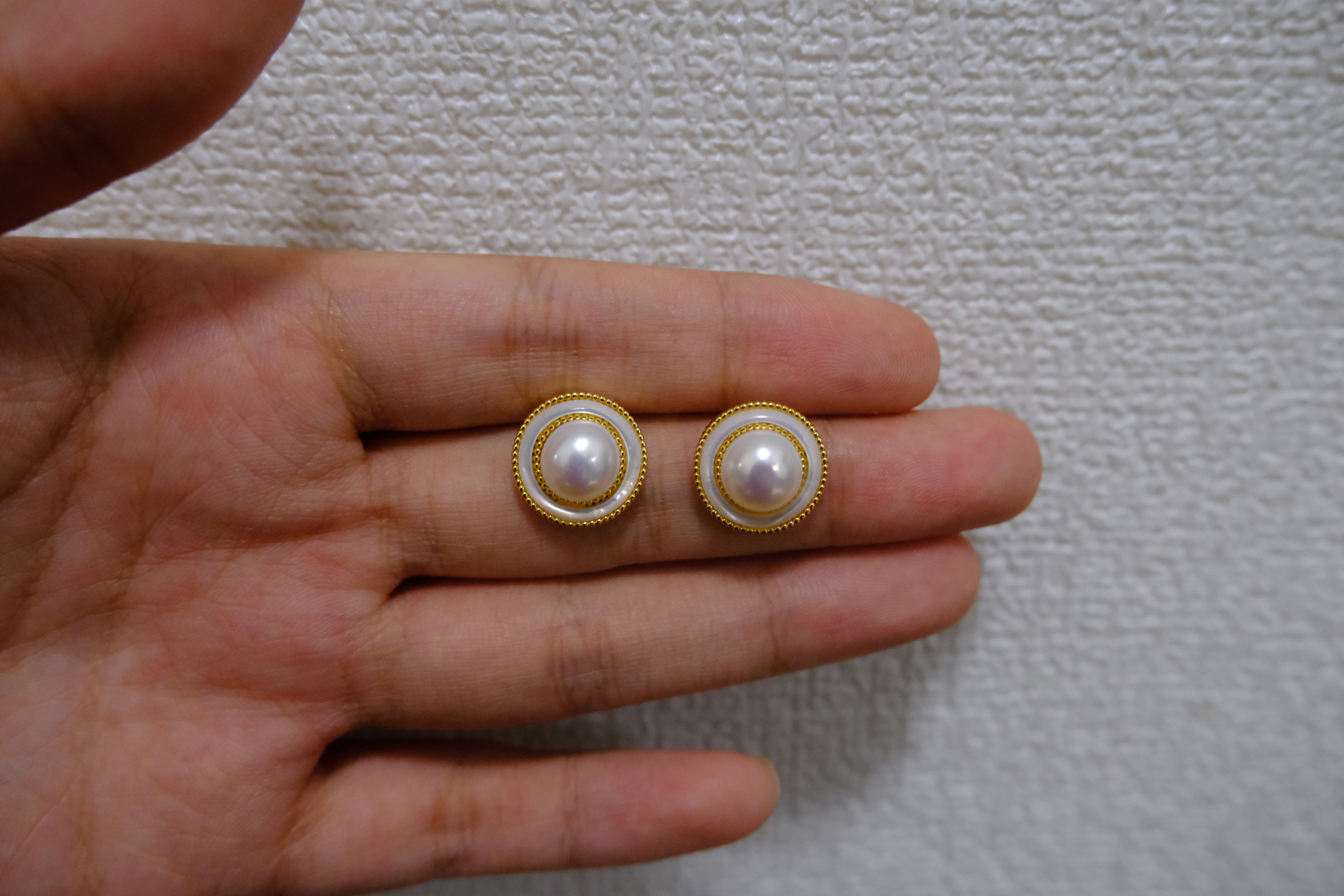 Two round freshwater pearl earrings with gold-plated frames are held in a hand.