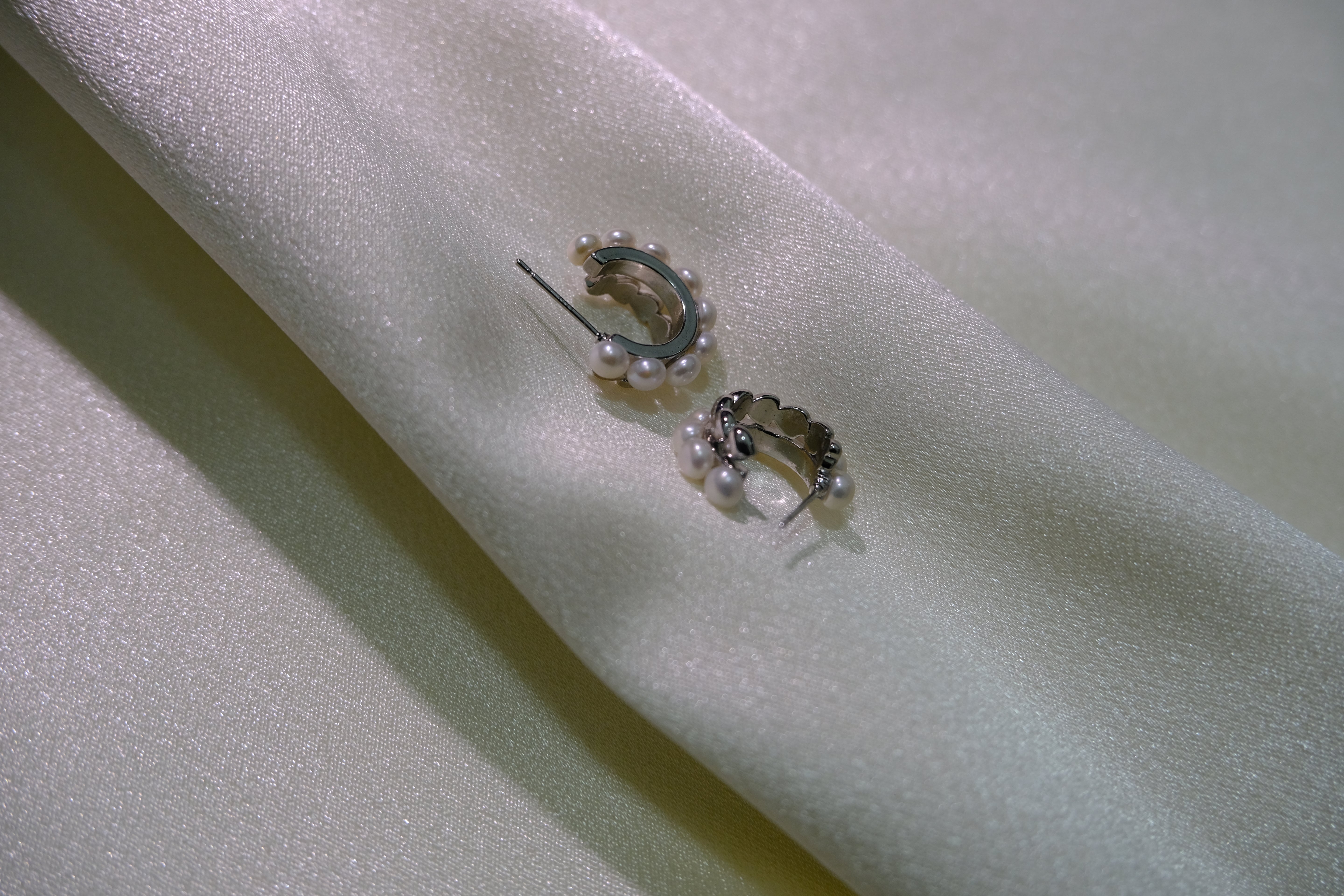 Two silver half-hoop earrings with small pearls are displayed on a light fabric.