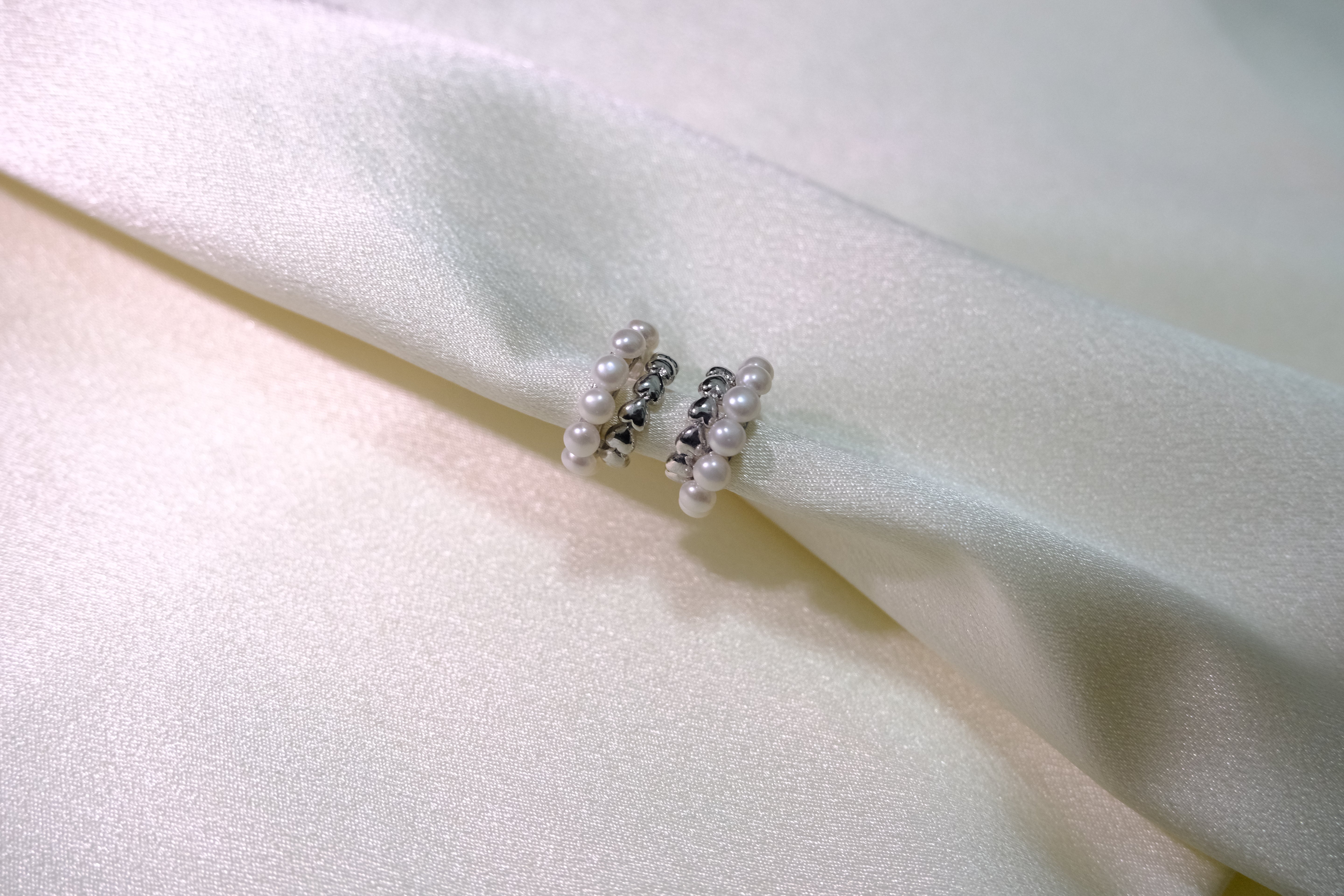 Two silver half-hoop earrings with small pearls are displayed on a white fabric.