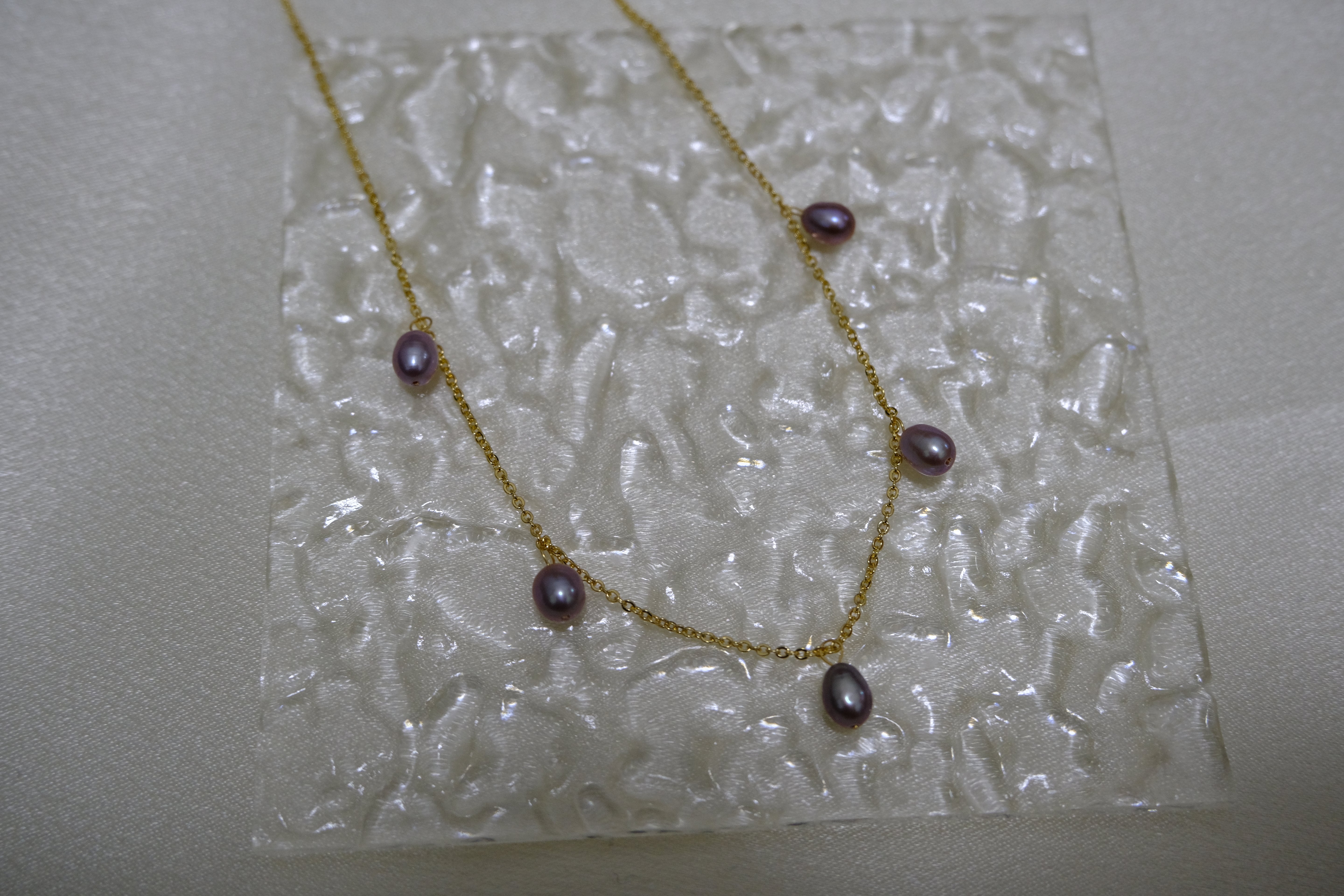 Dainty gold-plated necklace with five purple freshwater pearls on a textured white surface.