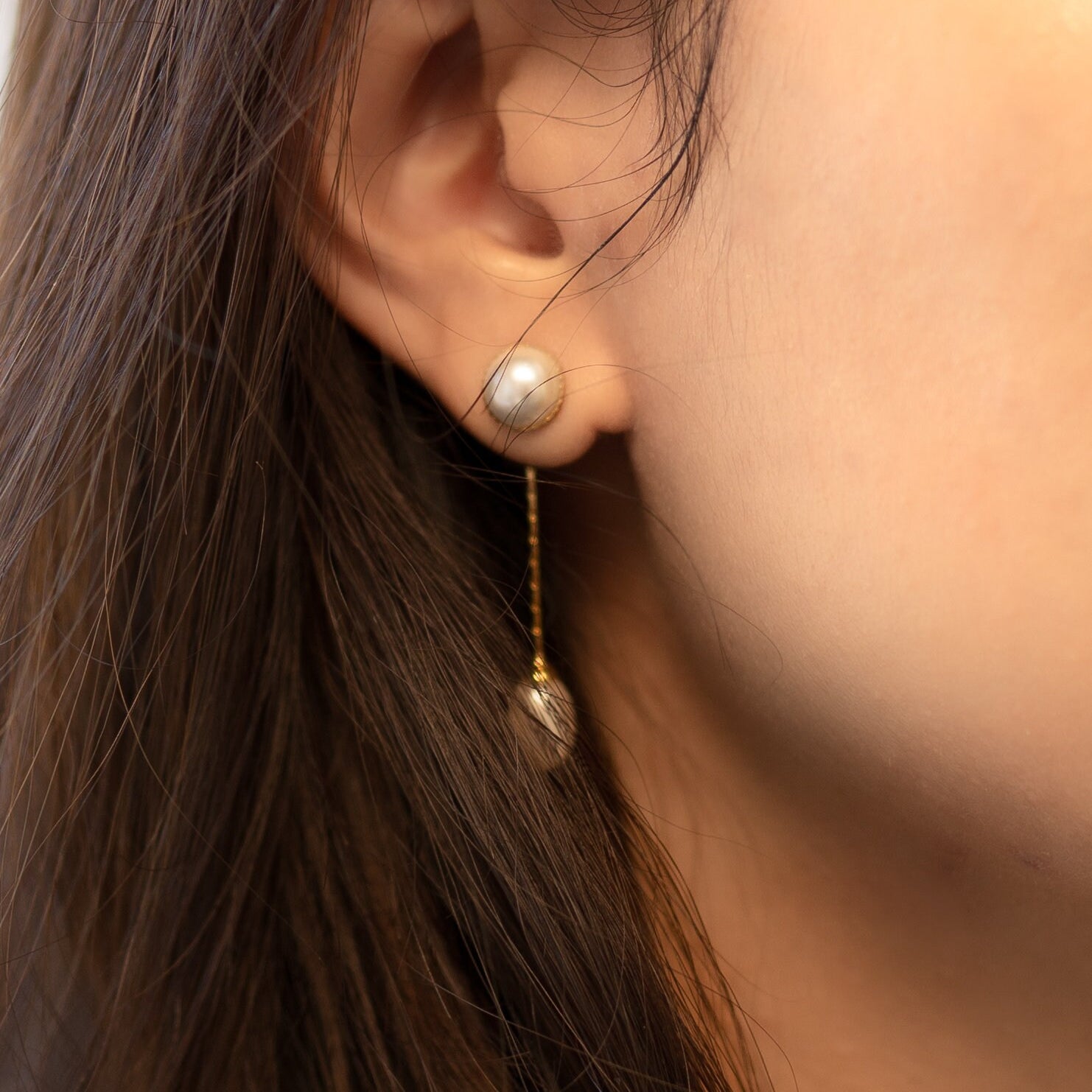 Elegant pearl dangle earrings with a gold-plated chain are worn on an ear.
