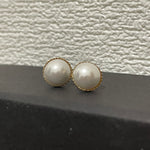 Two round pearl stud earrings with gold-toned settings rest on a dark surface.