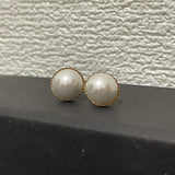 Two round pearl stud earrings with gold-toned settings rest on a dark surface.