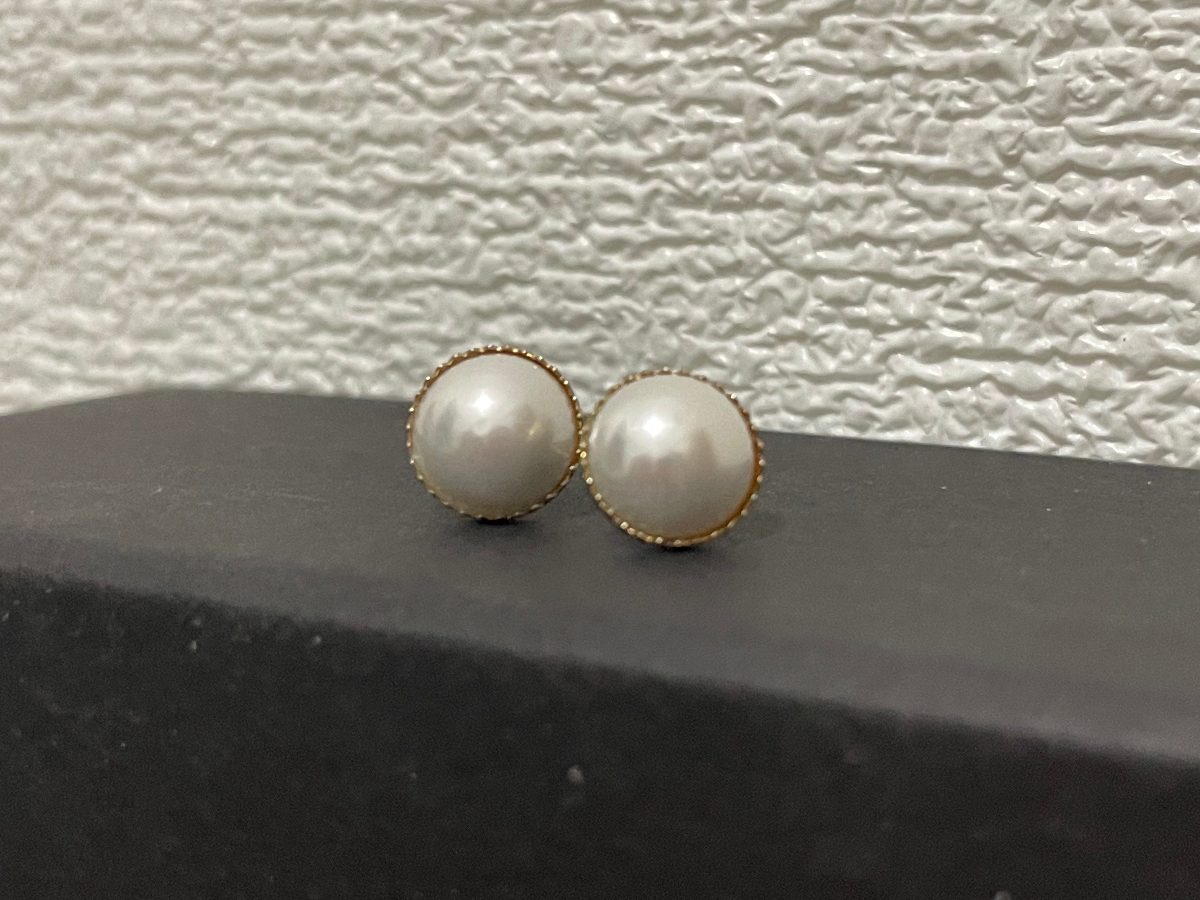 Two round pearl stud earrings with gold-toned settings rest on a dark surface.