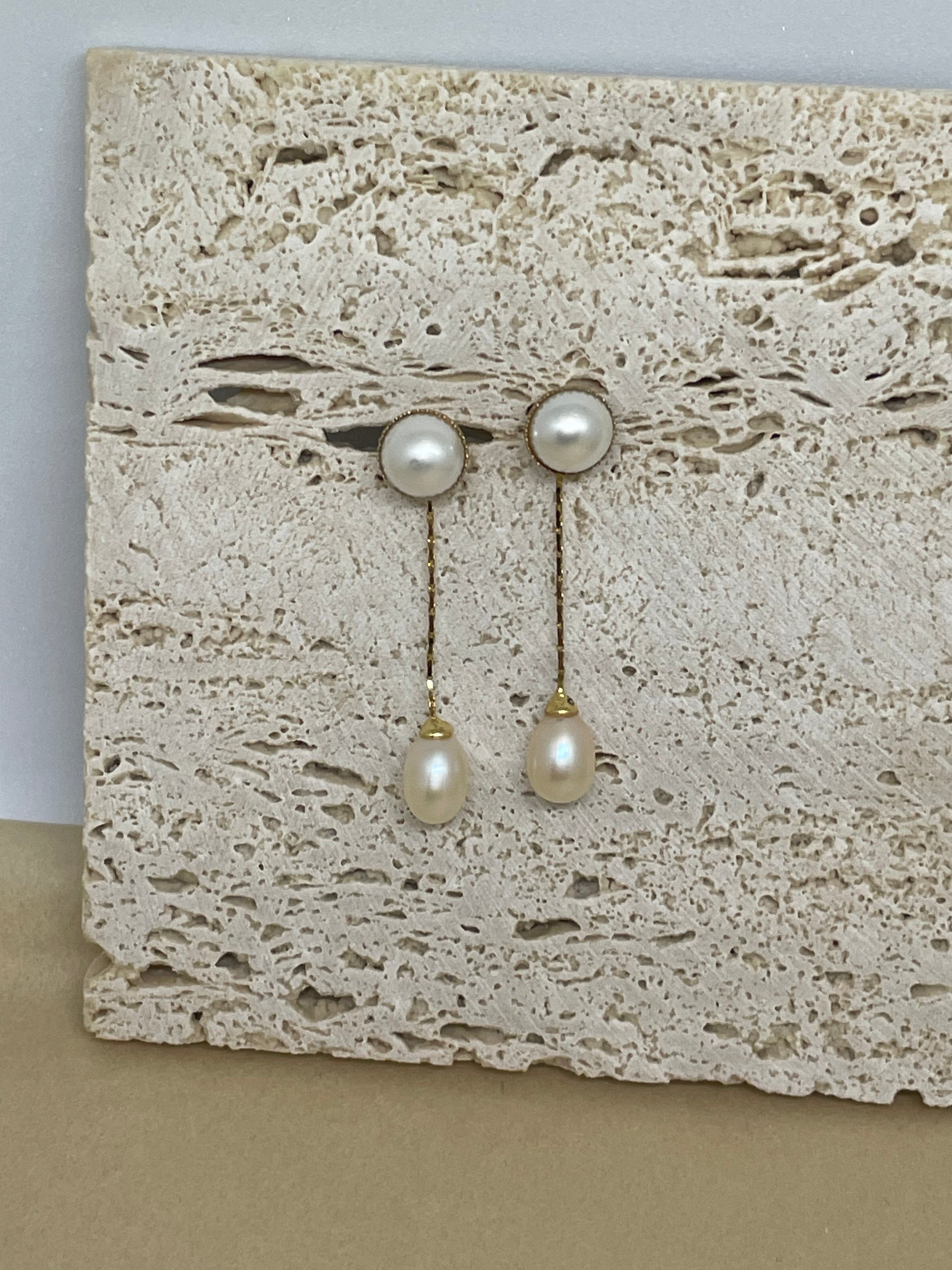 Elegant pearl dangle earrings with gold-plated chains on a textured stone surface.