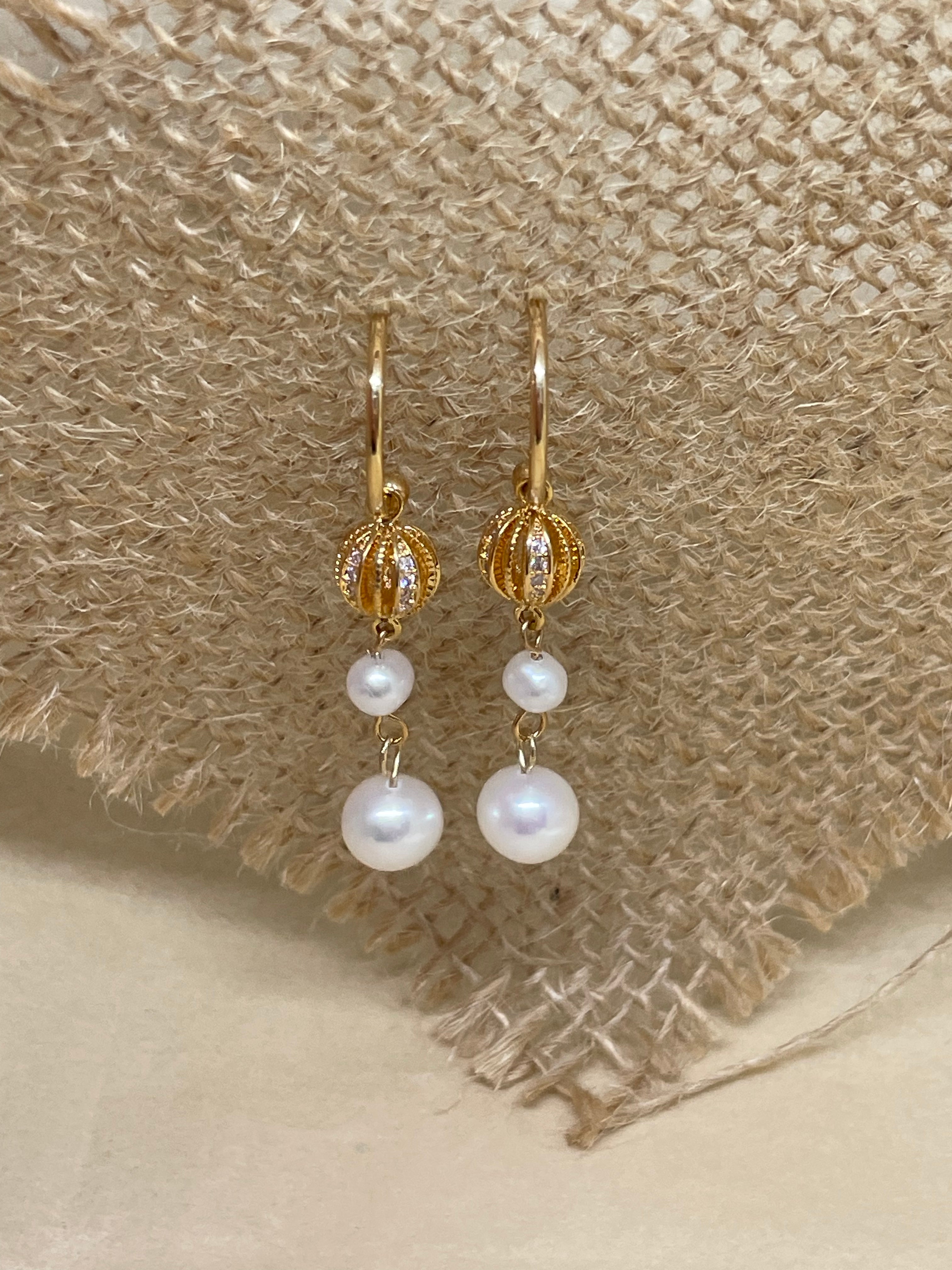 Gold hoop earrings with a textured gold bead and two white pearls on a burlap background.