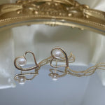 Gold heart-shaped dangle earrings with a single pearl and delicate chains, reflected on a mirror.