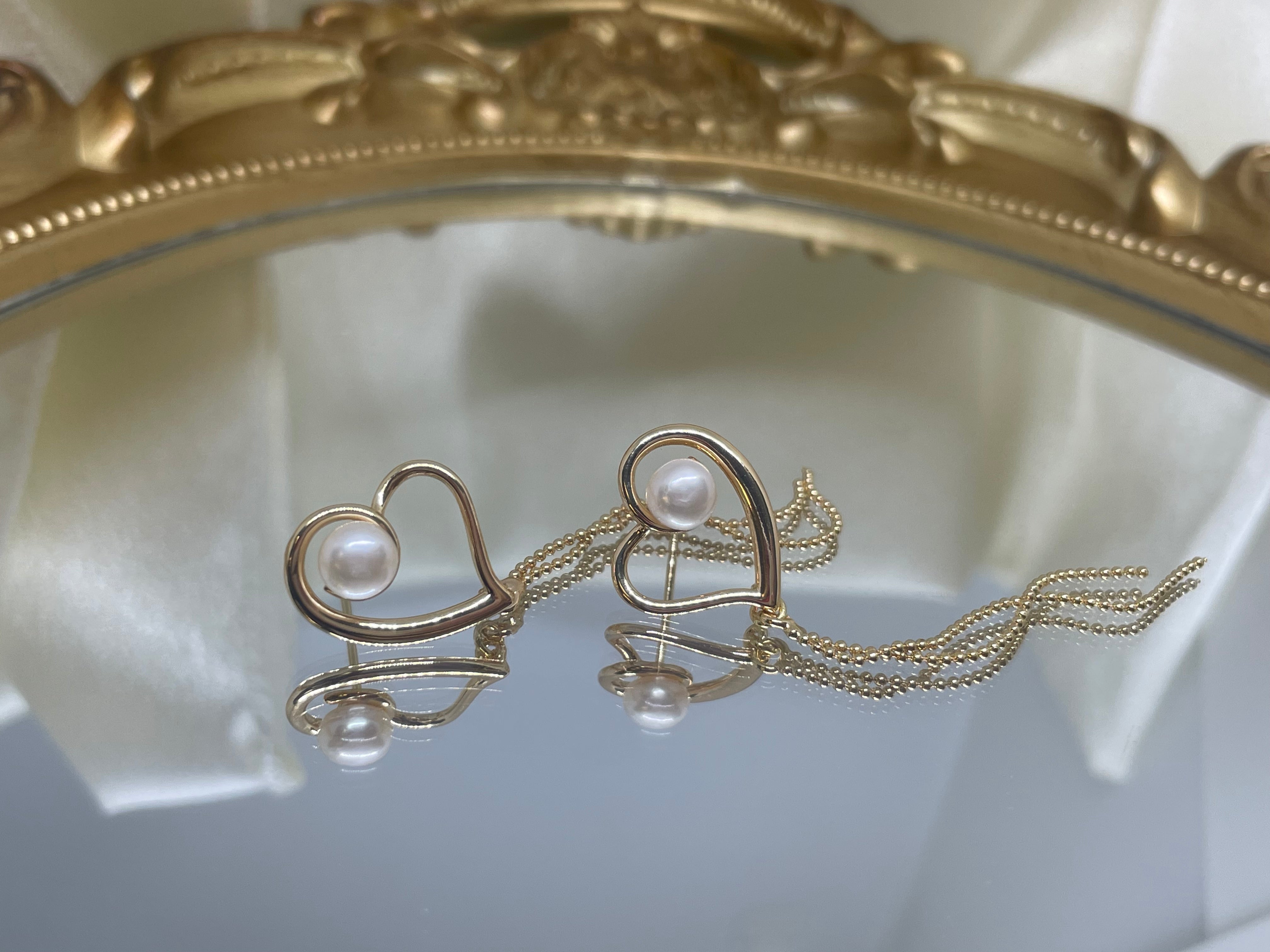 Gold heart-shaped dangle earrings with a single pearl and delicate chains, reflected on a mirror.