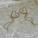 Two gold heart-shaped dangle earrings with freshwater pearls on a textured surface.