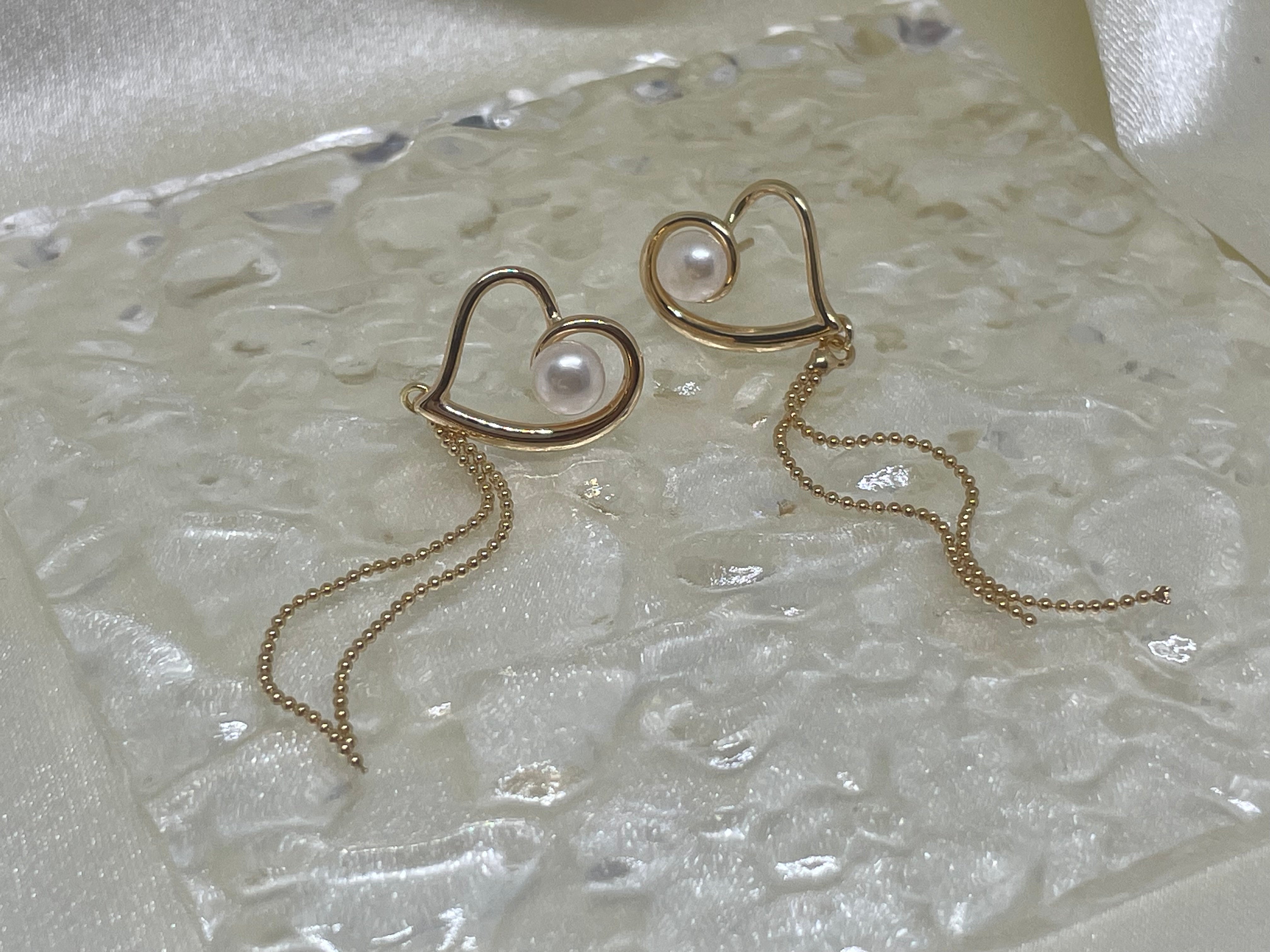 Two gold heart-shaped dangle earrings with freshwater pearls on a textured surface.