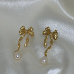 Two gold bow earrings with dangling freshwater pearls on a white satin background.