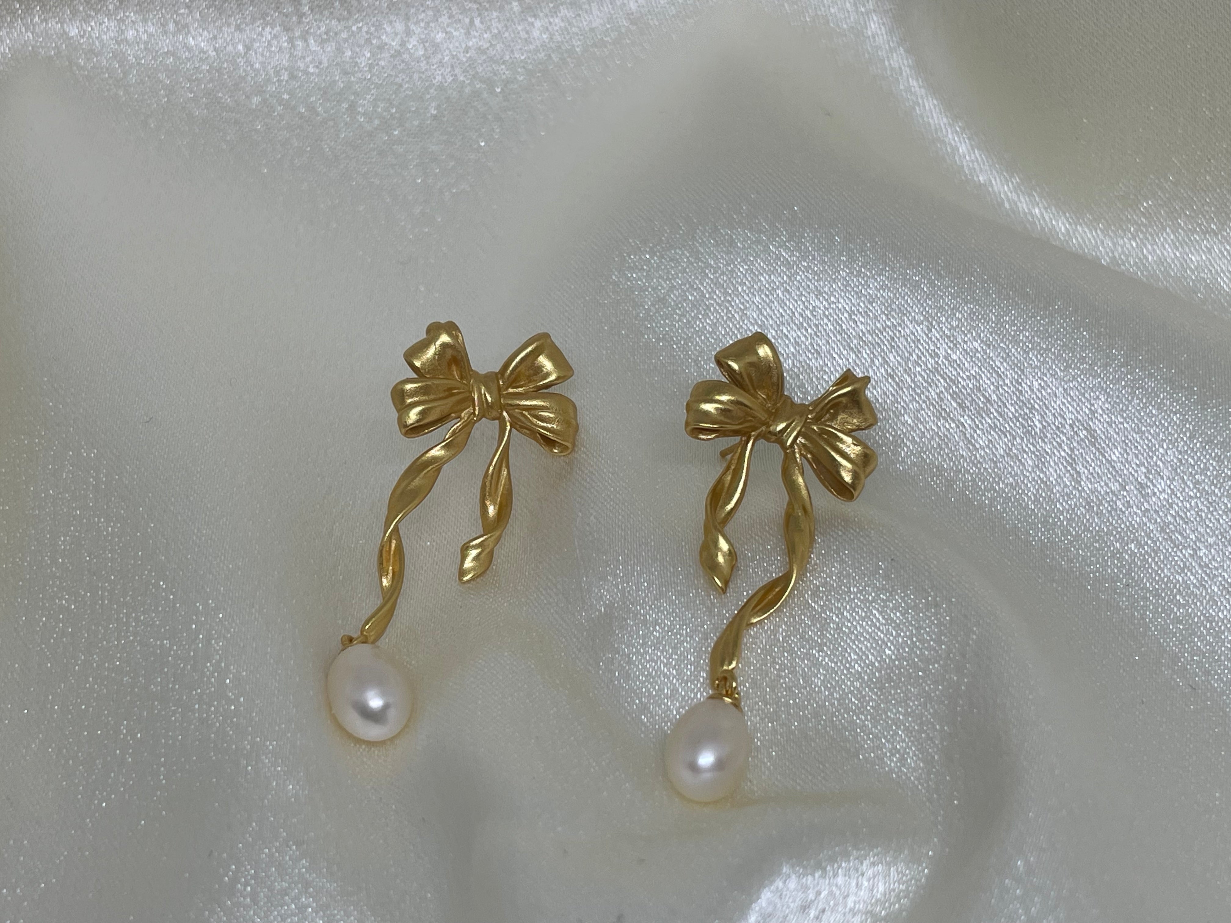 Two gold bow earrings with dangling freshwater pearls on a white satin background.