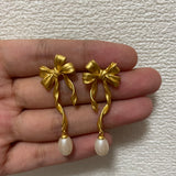 Freshwater pearl earrings with a gold bow design are held in a hand.