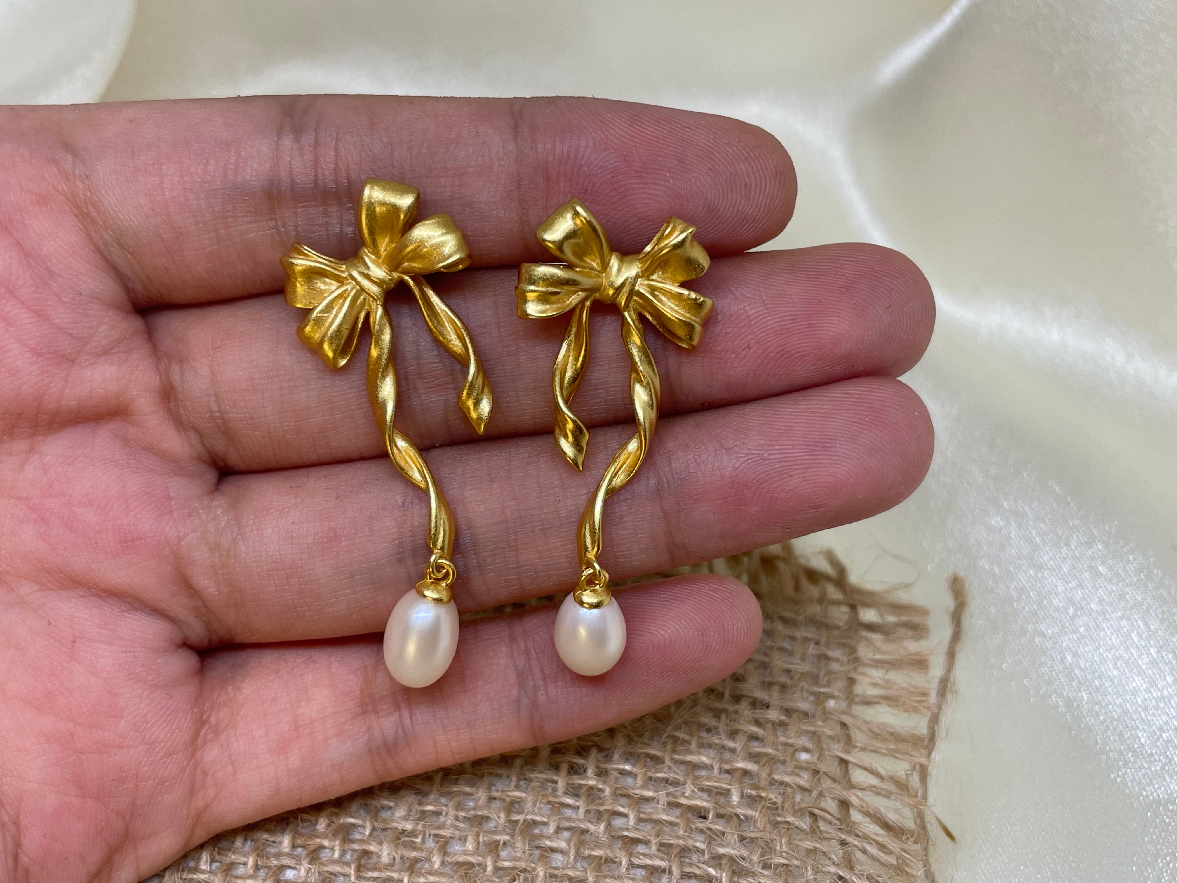 Gold bow earrings with dangling freshwater pearls held in a person's hand.
