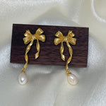 Gold bow earrings with dangling freshwater pearls on a dark wood block.