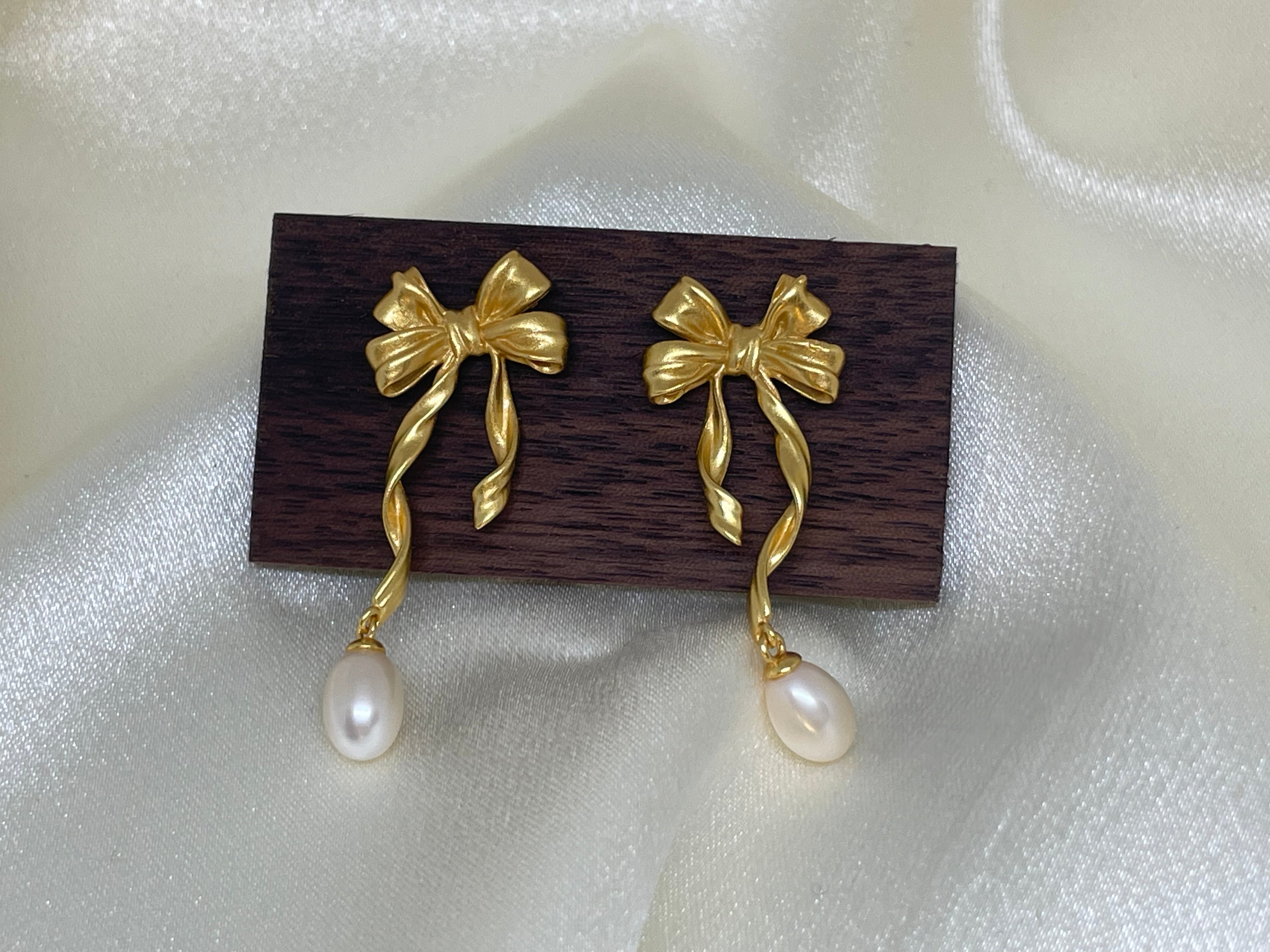 Gold bow earrings with dangling freshwater pearls on a dark wood block.
