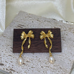 Freshwater pearl earrings with a gold bow and dangling pearl are displayed on wood.