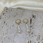 Two gold hoop earrings with three dangling white pearls each, on a textured white surface.