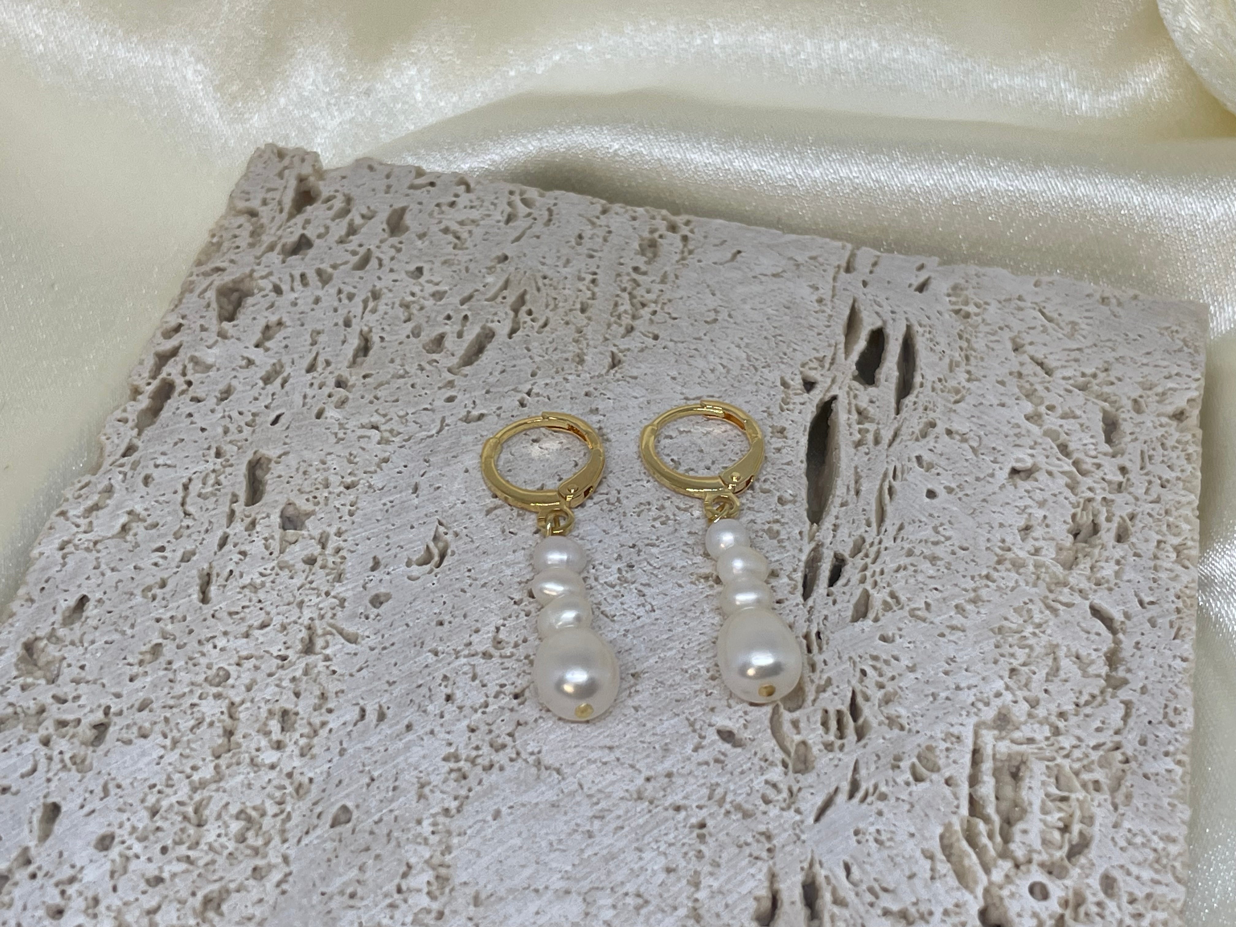 Two gold hoop earrings with three dangling white pearls each, on a textured white surface.