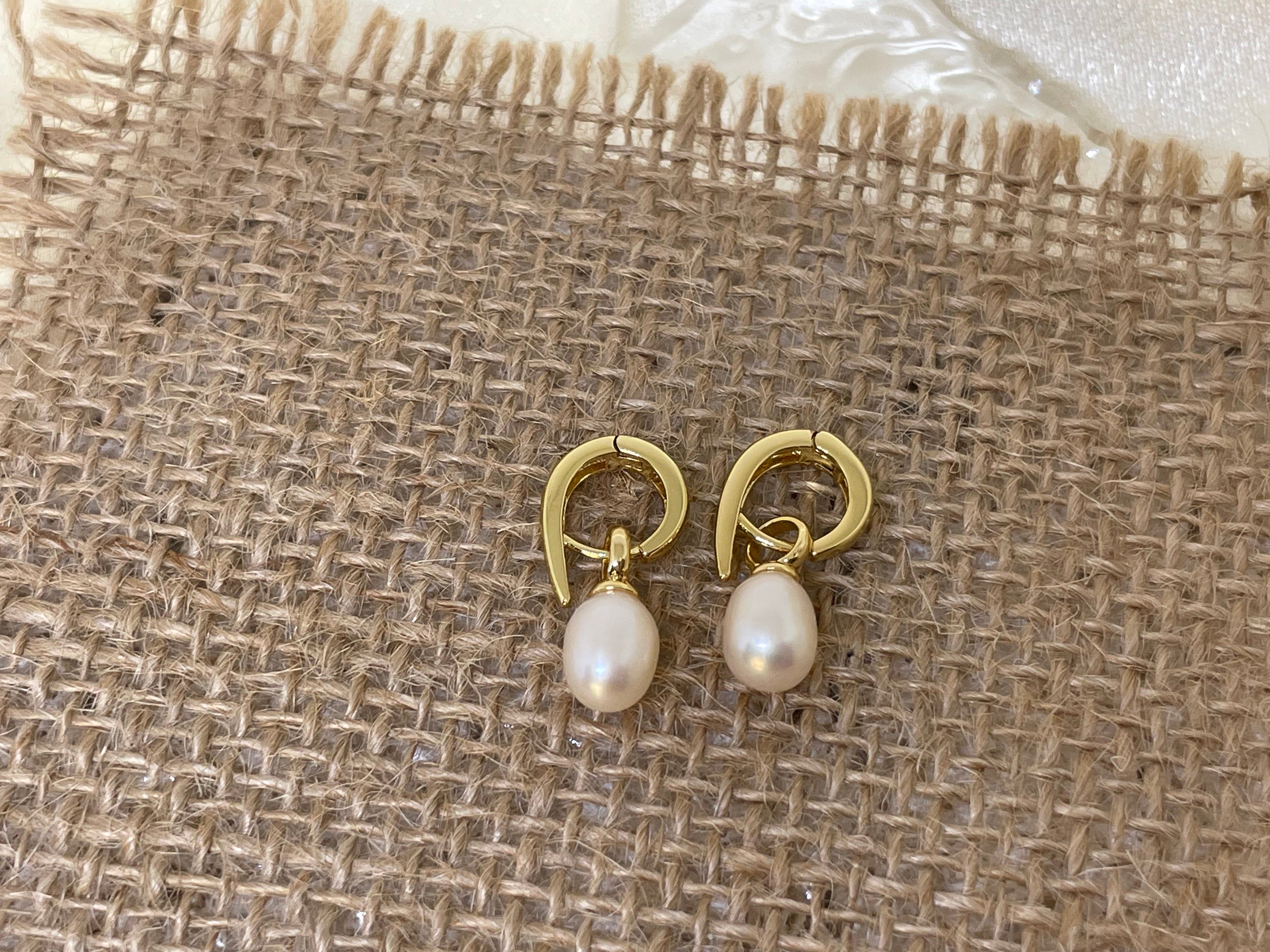 Two gold twist earrings with freshwater pearls are displayed on a burlap surface.