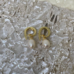 A pair of gold twist earrings with dangling freshwater pearls rests on a textured surface.