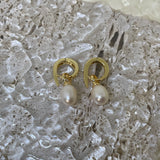 A pair of gold twist earrings with dangling freshwater pearls rests on a textured surface.