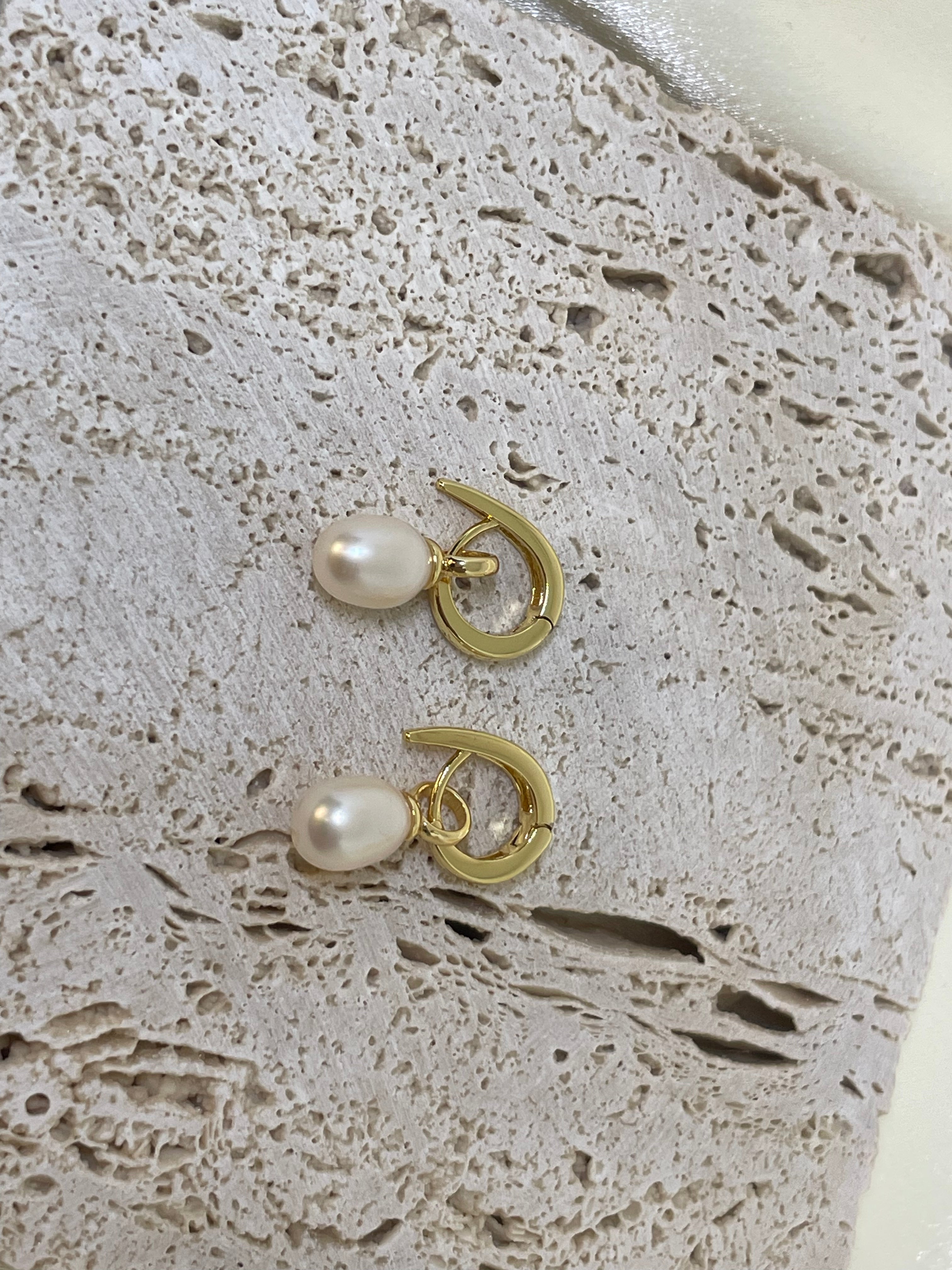 Two gold twist earrings with freshwater pearls are displayed on a textured surface.
