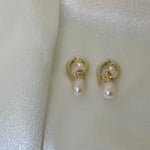 Two gold twist earrings with freshwater pearl drops are displayed on a white, shimmery fabric.